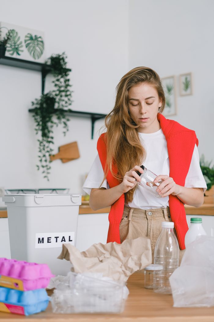 Serious female standing at table with used plastic bags and bottles near egg boxes and paper while taking off lid from glass jar sorting trash